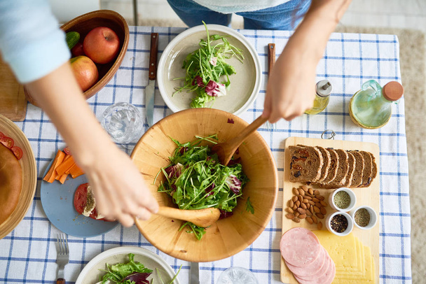 Vista superior de primer plano de las manos femeninas mezclando ensalada verde en un tazón de madera en la mesa con comida saludable mientras prepara la cena familiar
 - Foto, Imagen