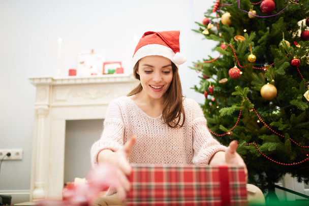 Joyful pretty woman wearing knitted sweater and Santa hat unwrapping Christmas gift box with interest while sitting by fireplace, portrait shot - Photo, Image