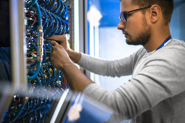 Portrait latéral de jeune homme portant des lunettes connectant des câbles dans une armoire serveur tout en travaillant avec un superordinateur dans un centre de données
 - Photo, image