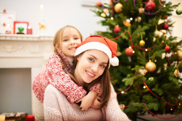 Family portrait of attractive woman wearing Santa hat sitting by fireplace while her cute little daughter embracing her tenderly, decorated Christmas tree behind  them - Photo, Image
