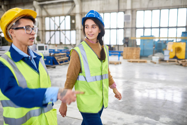 Confident experienced forewoman walking with female colleague over factory workshop and explaining her work - 写真・画像