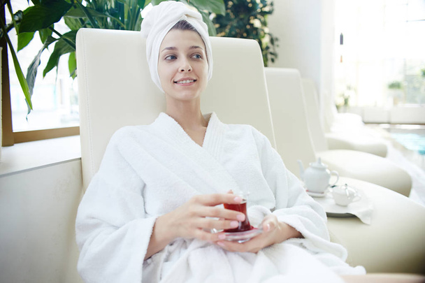 Портрет красивой молодой женщины, улыбающейся, расслабляясь в шезлонге у бассейна в халате и полотенце на голове
 - Фото, изображение