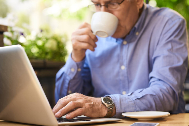 Closeup image of senior man using laptop computer enjoying coffee break in outdoor cafe lounge - Photo, image