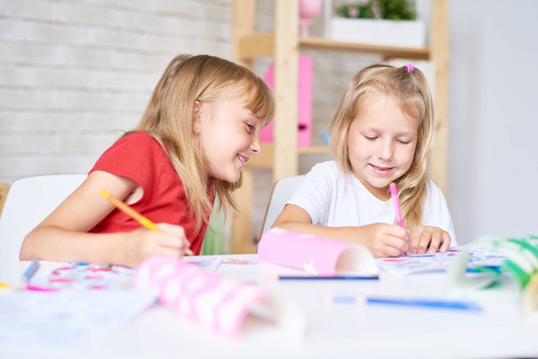 Portrait de deux jolies petites sœurs coloriant des images ensemble assis à table dans la salle de jeux et souriant
 - Photo, image