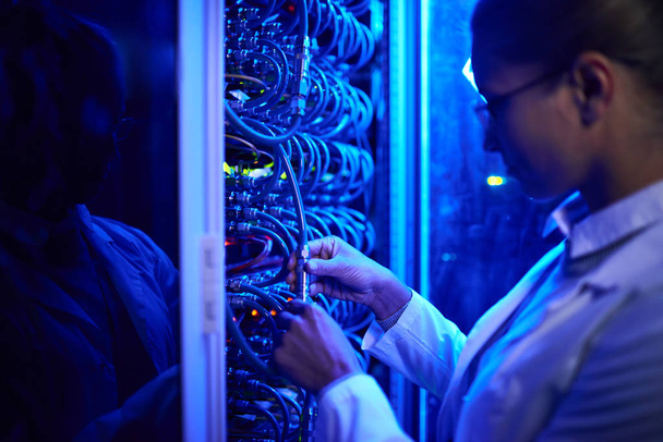 Retrato de vista lateral de una mujer joven que usa bata de laboratorio conectando cables en el gabinete del servidor mientras trabaja con supercomputadora en luz de neón
 - Foto, Imagen