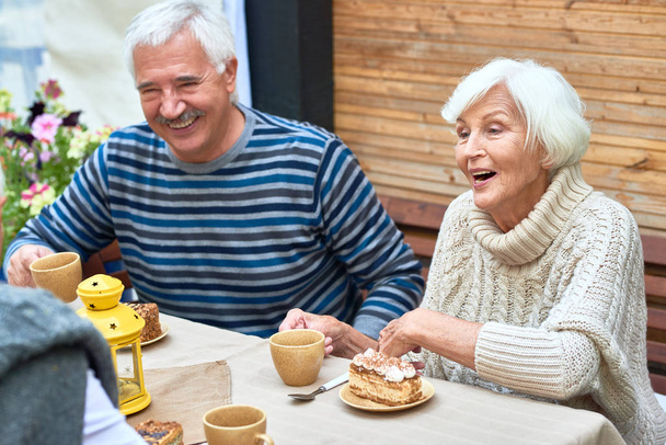 Портрет счастливой пожилой пары, смеющейся, наслаждающейся ужином с друзьями и семьей, пьющей чай с тортами на открытой террасе
 - Фото, изображение