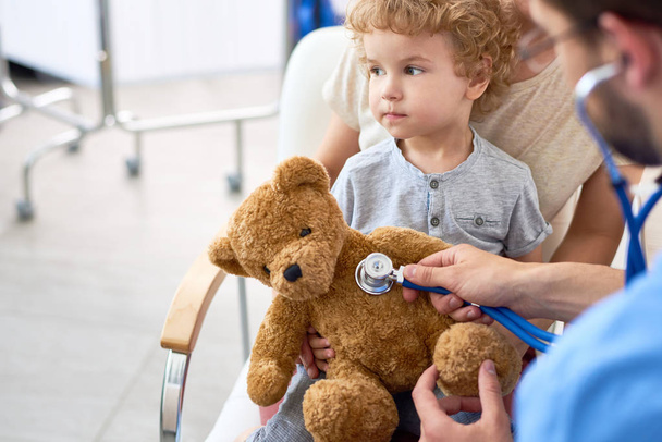 Porträt eines entzückenden lockigen Kindes in der Arztpraxis mit Teddybär-Spielzeug, bei dem der Kinderarzt mittels Stethoskop dem Herzschlag zuhört - Foto, Bild