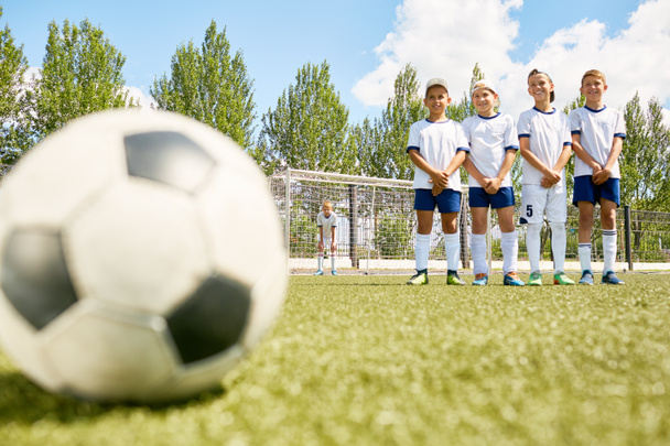 Портрет щасливих хлопчиків у юнацькій футбольній команді, що стоїть поспіль перед тренуванням з футбольним м'ячем, що лежить на траві на передньому плані
 - Фото, зображення