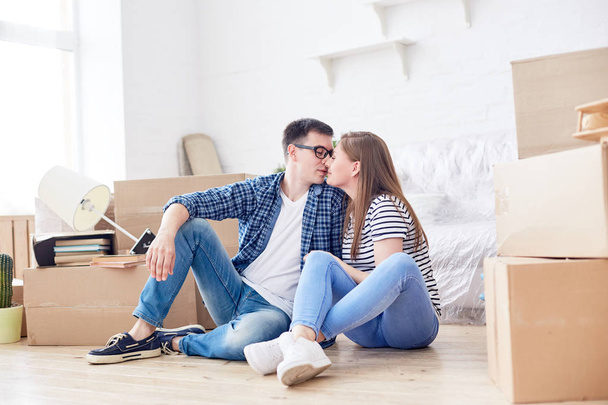 Retrato de larga duración de pareja joven y cariñosa expresando su sensación de beso mientras se sienta en el suelo de la sala de estar rodeada de cajas móviles
 - Foto, Imagen