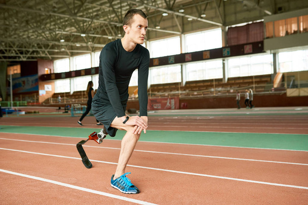 Portrait of determined amputee sportsman warming up before running practice in modern gym stretching legs - Photo, Image