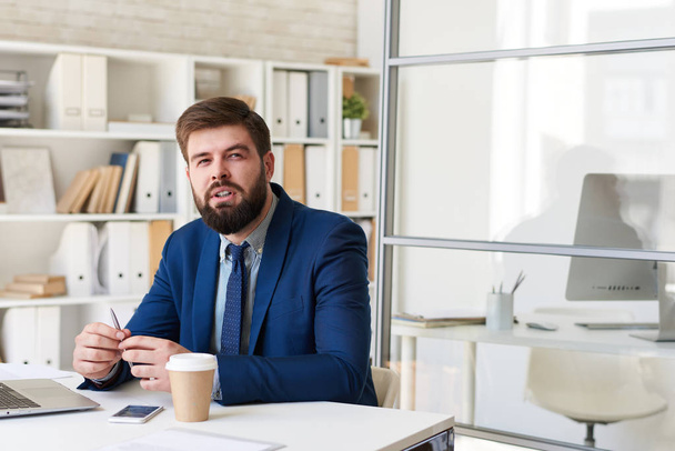 Портрет успешного бородатого бизнесмена, задумчиво смотрящего в сторону, работающего за столом в современном офисе
 - Фото, изображение