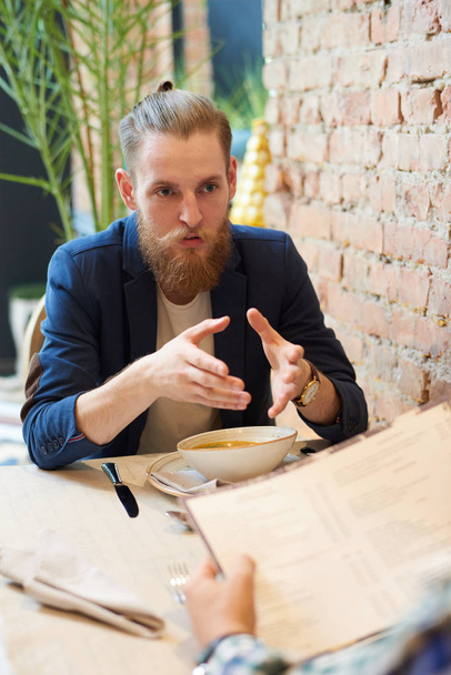 Portrait d'un bel homme barbu expliquant quelque chose à un collègue assis à table dans un café pendant la pause déjeuner
 - Photo, image