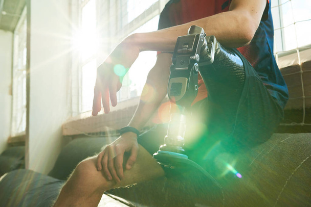 Primer plano del deportista amputado sentado en el banco en el gimnasio moderno, se centran en la pierna protésica a la luz del sol con destello de lente
 - Foto, Imagen