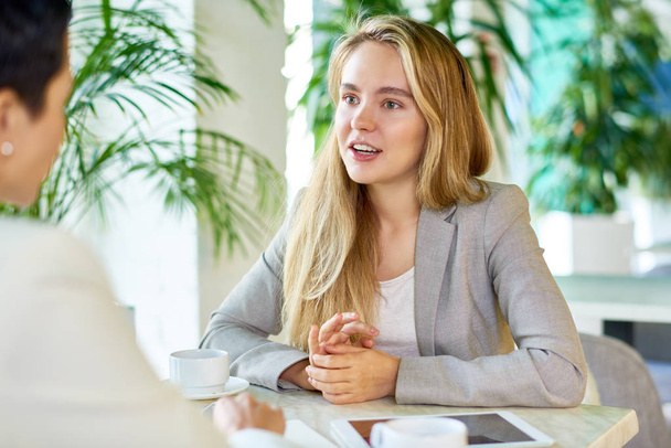 Portrait of two modern young women discussing work sitting at table in cafe during business meeting, focus on young blond trainee talking enthusiastically - Photo, Image