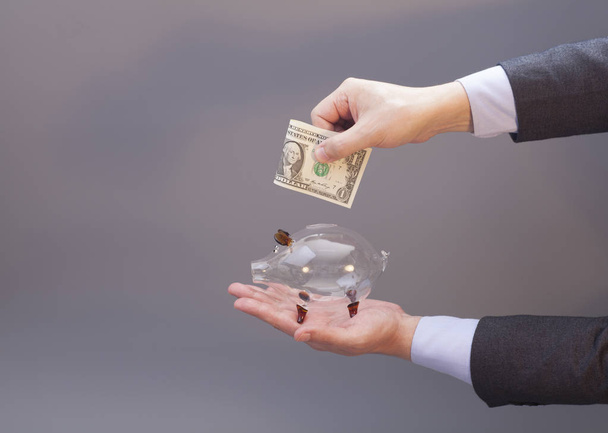 Businessman putting one dollar bill into glass piggy bank on gray - Fotografie, Obrázek