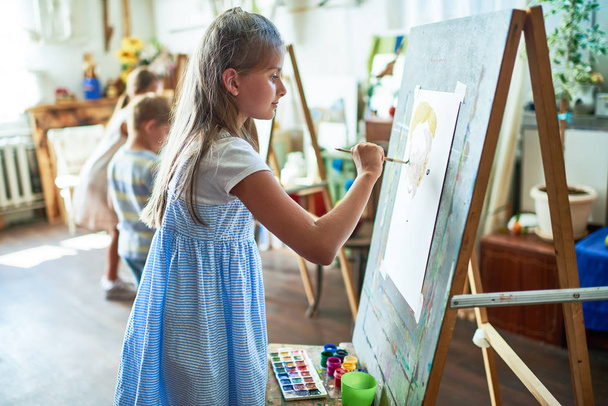Kindergruppe malt auf Staffeleien während des Kunstunterrichts im sonnenbeschienenen Atelier, hübsche Schülerin im Vordergrund - Foto, Bild
