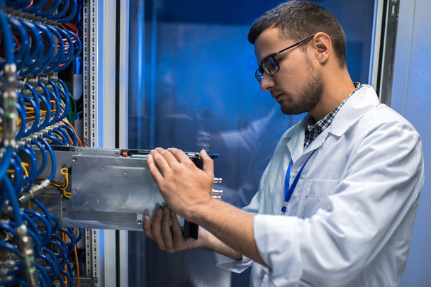 Retrato de vista lateral del joven con bata de laboratorio sacando el servidor blade del gabinete mientras trabaja con supercomputadora
 - Foto, Imagen