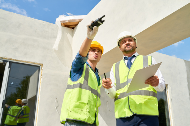Low angle view of confident bearded businessman wearing suit and hardhat talking to young foreman while checking construction phase of building - Photo, Image