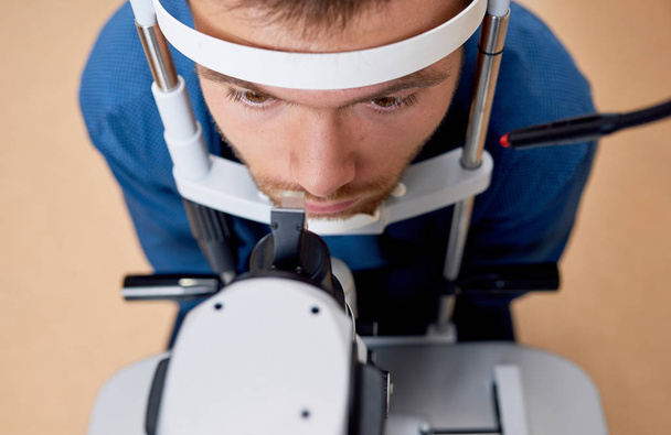 Primer plano de ángulo alto del hombre joven mirando la máquina de lámpara de hendidura, apoyando la cabeza en el soporte durante las pruebas de visión en la oficina del optometrista
 - Foto, Imagen