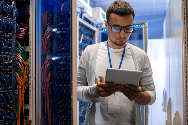Portrait of modern young man using digital tablet standing by server cabinet while working with supercomputer in blue light - Photo, Image