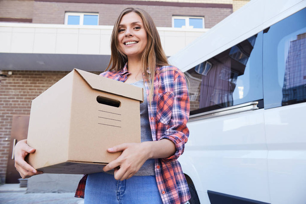 Porträt einer glücklichen jungen Frau, die einen Karton in der Hand hält und neben einem fahrenden Lieferwagen in die Kamera lächelt - Foto, Bild