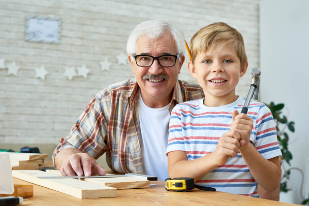 Portret szczęśliwy człowiek starszy pozowanie z małym wnukiem patrząc na kamery i uśmiechając się podczas pracy z drewna razem w domu - Zdjęcie, obraz