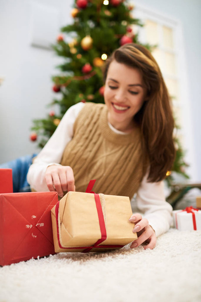 Porträt einer attraktiven Frau mit langen dunklen Haaren, die auf einem gemütlichen Teppich liegt und das Weihnachtsgeschenk öffnet, geschmückter Weihnachtsbaum hinter sich - Foto, Bild