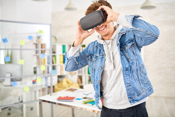Retrato de un joven disfrutando de un vídeo en gafas VR sonriendo de pie en un espacio de oficina moderno
 - Foto, imagen