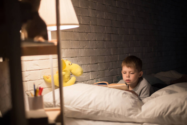 Retrato de lindo niño leyendo cuento de hadas en la cama bajo la luz de la lámpara, libro de apoyo en la almohada en la habitación oscura por la noche
 - Foto, imagen