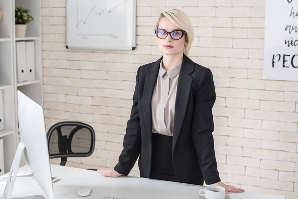 Portrait of blonde young businesswoman standing at desk in modern office, posing looking at camera with serious face expression - Photo, Image