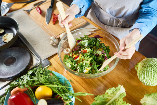Magas szög közelről, fiatal nő, a kötény keverés saláta üvegtál dolgozva, modern konyhában főzés - Fotó, kép