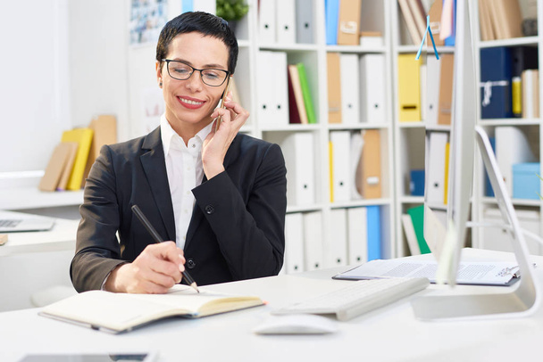 Attractive middle-aged designer writing down order details while talking to her client on mobile phone, interior of modern office on background - Fotografie, Obrázek