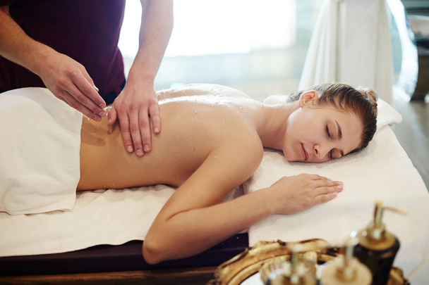 Porträt einer glückseligen jungen Frau, die eine Massage im Wellness-Center genießt, mit geschlossenen Augen auf einem Massagetisch liegt und lächelt, während sich der männliche Masseur ihren Rücken reibt - Foto, Bild