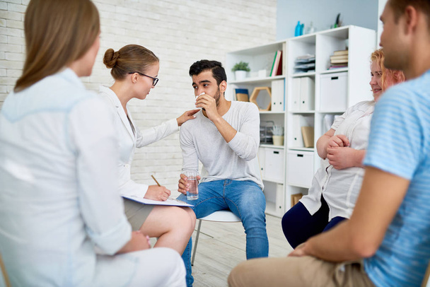 Портрет женщины-психиатра, утешающей плачущего молодого человека на сеансе групповой терапии с другими пациентами, сидящими в кругу вокруг них
 - Фото, изображение