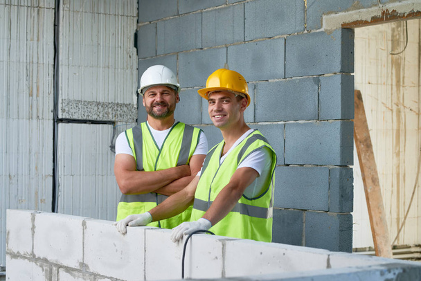 Retrato de grupo de trabajadores de la construcción alegres mirando a la cámara con sonrisas de dientes blanco tomando un breve descanso del trabajo, interior del edificio sin terminar en el fondo
 - Foto, imagen