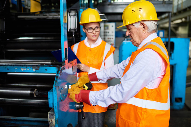 Retrato de una mujer joven observando máquinas operativas de trabajadores de fábrica senior, ambas con sombreros duros y chaquetas reflectantes
 - Foto, Imagen