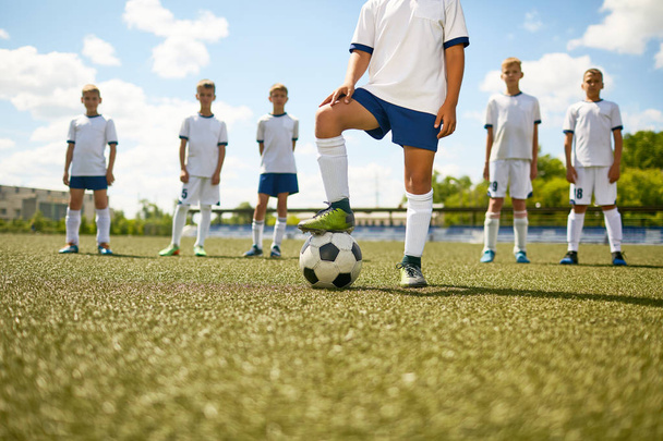 Retrato de seção baixa de capitão de futebol em pé no centro de campo pisando na bola com o resto da equipe em segundo plano
 - Foto, Imagem
