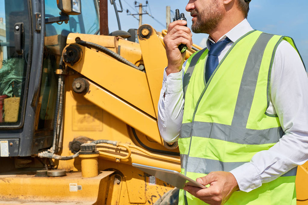 Closeup of construction worker wearing suit and reflective jacket talking via portable radio set standing by road roller and using digital tablet - Photo, Image