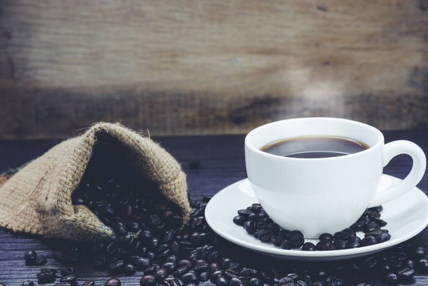 Боковой вид Кофейная чашка с дымом и кофейными зёрнами в мешке на деревянном столе, Напитки и расслабляющая концепция, для рекламы
 - Фото, изображение