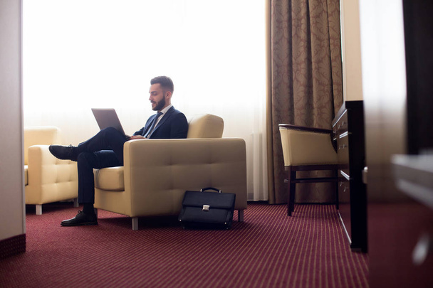 Po celé délce portrét pohledný mladý podnikatel používat přenosný počítač v hotelovém pokoji, relaxační křeslo, kopie prostor - Fotografie, Obrázek