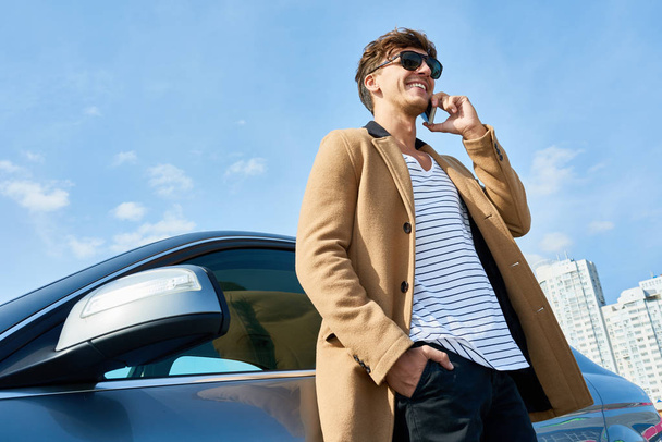 Low angle portrait of handsome young man speaking by phone standing in sunlight leaning on car outdoors against clear blue sky, copy space - Photo, image