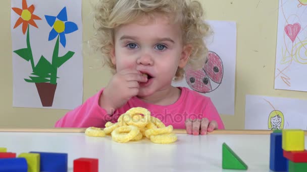 Χαριτωμένο πρόσωπο κορίτσι τρώει καλαμπόκι κύκλο κάθεται κοντά της επιτραπέζια παιχνίδια και τα σχέδια - Πλάνα, βίντεο
