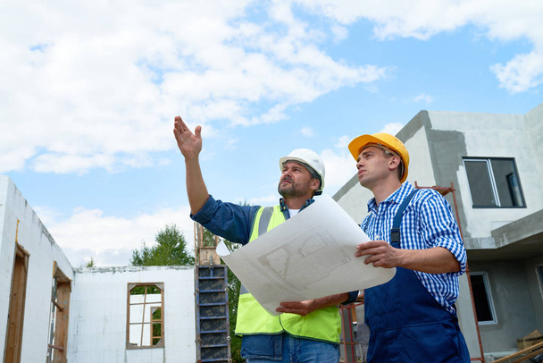 Retrato de dos trabajadores de la construcción con sombreros discutiendo los planos en el sitio, señalando hacia arriba
 - Foto, Imagen