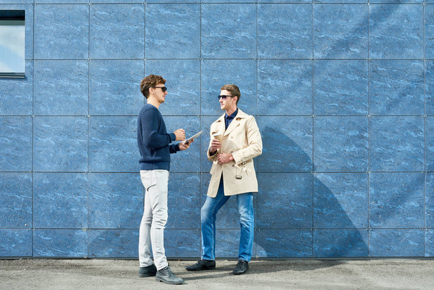 Täyspitkä muotokuva kahdesta menestyneestä nuoresta liikemiehestä juttelemassa ulkona kahvitauon aikana seisomassa betoniseinää vasten, kopioimassa
 - Valokuva, kuva