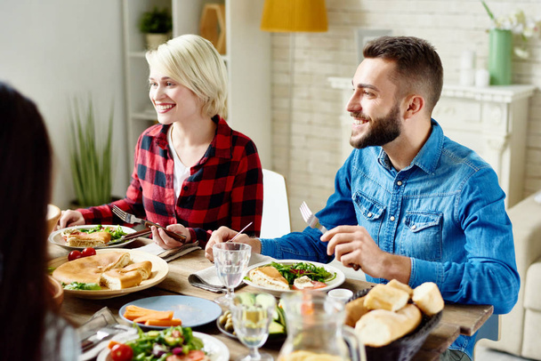 Portrait of happy young man and woman sitting at table with delicious meals, enjoying festive dinner with friends at home gathering - Photo, Image