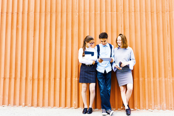 Retrato de tres niños de la escuela de pie contra la pared naranja brillante usando tableta digital
 - Foto, Imagen