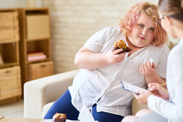 Portrait of obese young woman  eating cupcakes during therapy session with female psychiatrist - Photo, Image