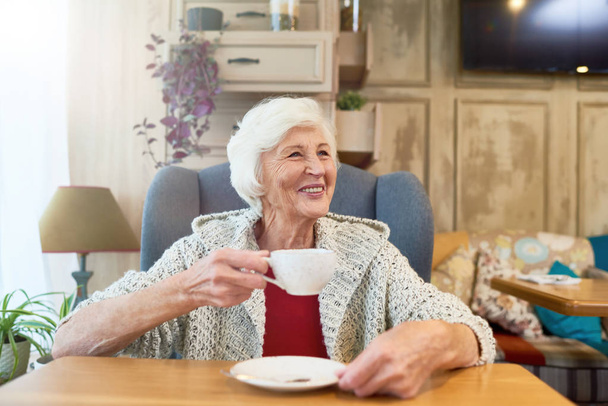 Muotokuva tyylikäs vanhempi nainen hymyilee onnellisesti katselee pois istuu iso mukava tuoli teekuppi
 - Valokuva, kuva