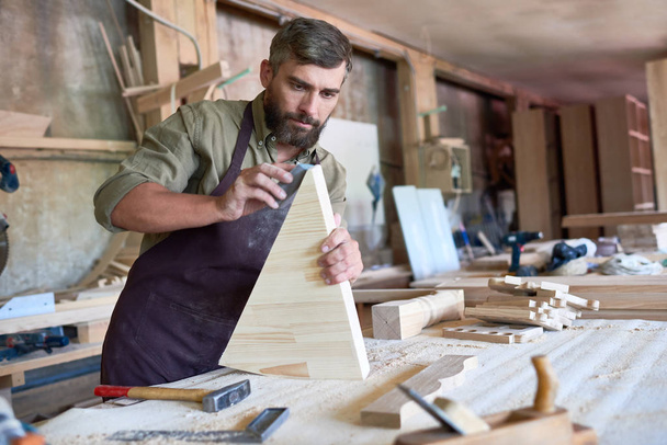 Portrait de charpentier barbu mature ponçant soigneusement la partie en bois dans un atelier moderne, espace de copie
 - Photo, image