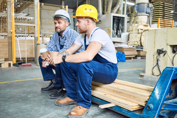 Full length portrait of two builders wearing hardhats taking break from work drinking coffee and resting sitting on pack of wood indoors - Photo, Image